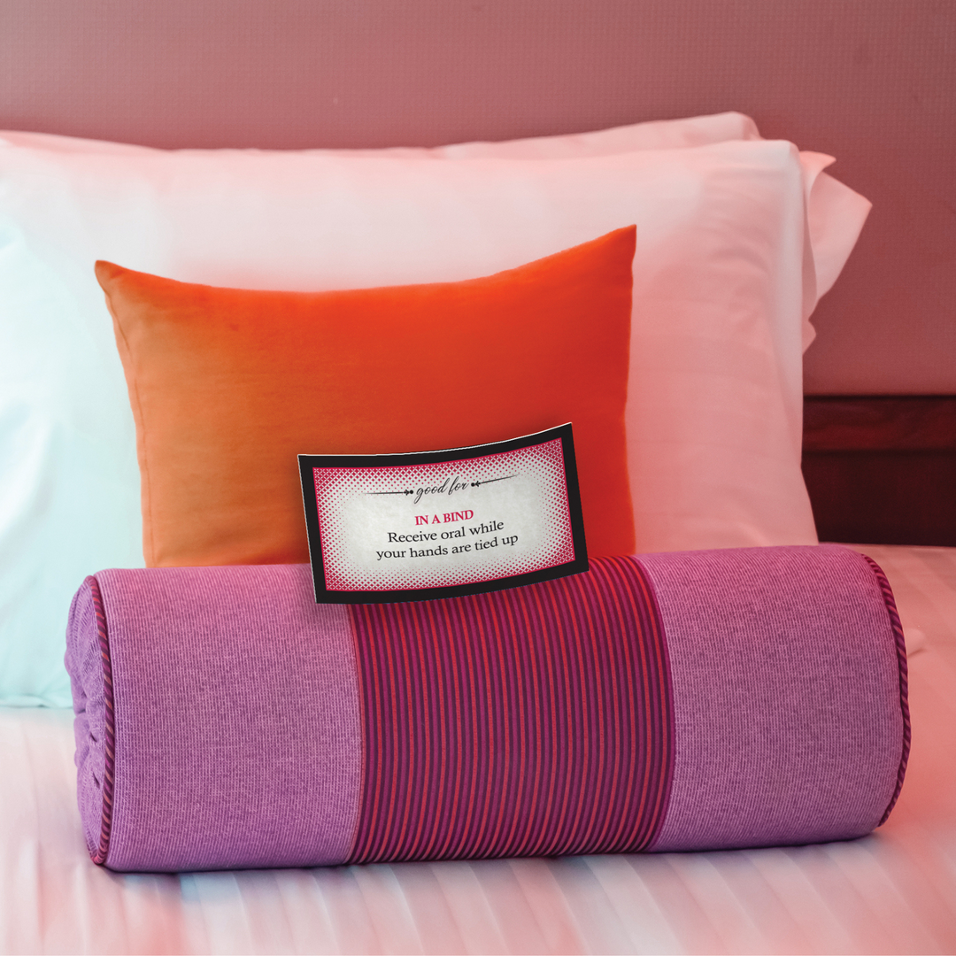 A card stating, "IN A BIND Receive oral while your hands are tied up.", laying on pink and orange pillows. 